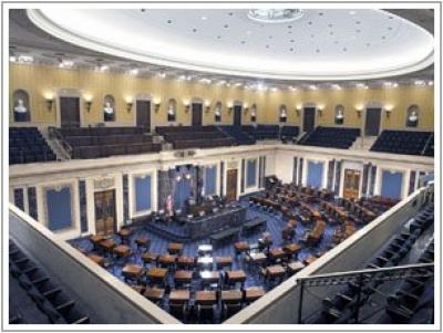 Senate chamber with a circular light in the ceiling shining above rows of chairs all facing a speakers podium in a half-circle shape